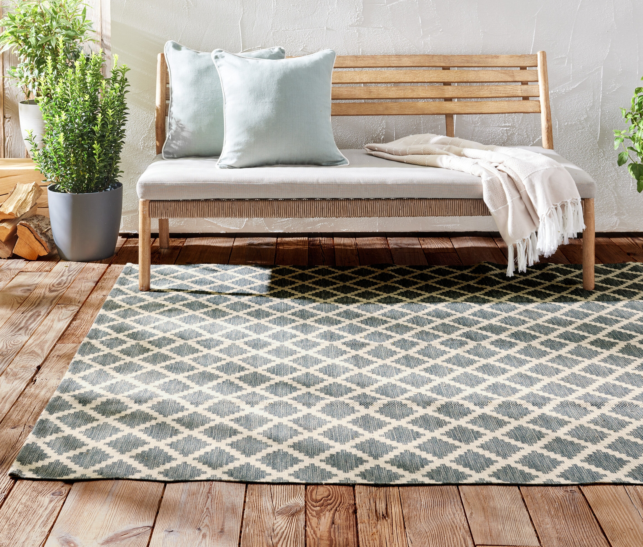 Sur un plancher en bois se trouve un tapis d’extérieur réversible d’environ 250 x 180 cm. Derrière, il y a un banc en bois avec des coussins et des plantes en pot.
