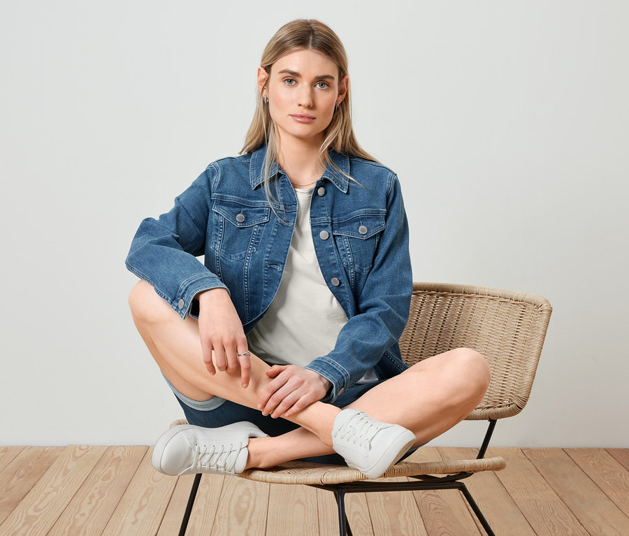 Une femme blonde est assise les jambes croisées sur une chaise. Elle porte une veste en jean, un t-shirt basique blanc et un bermuda en jean.