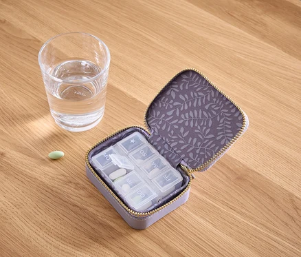 Sur une table en bois se trouvent un verre d'eau, une pilule verte et un pilulier violet ouvert contenant des pilules blanches.