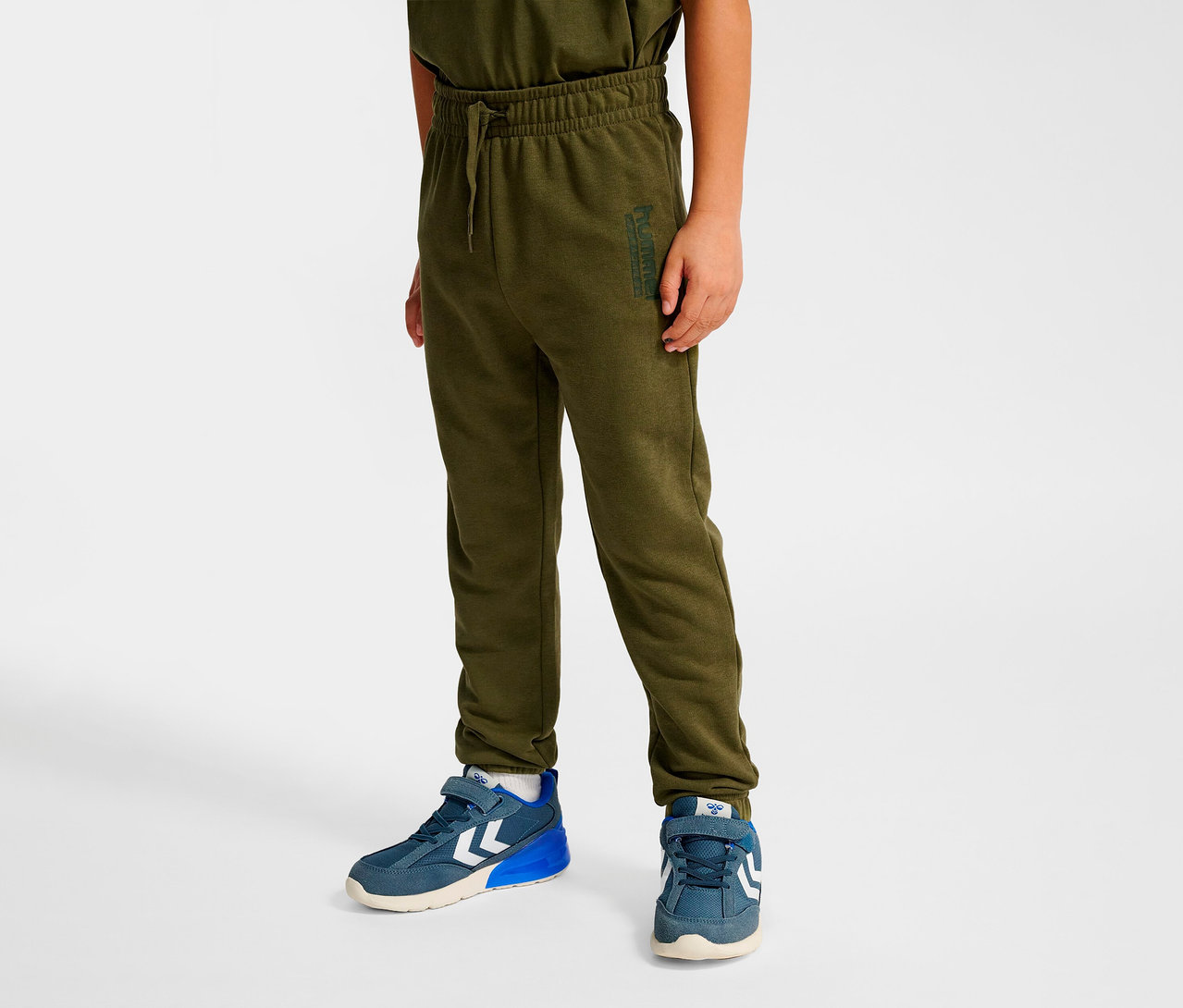 Un garçon porte un pantalon régulier HUMMEL HMLJR Base vert et des chaussures bleues.