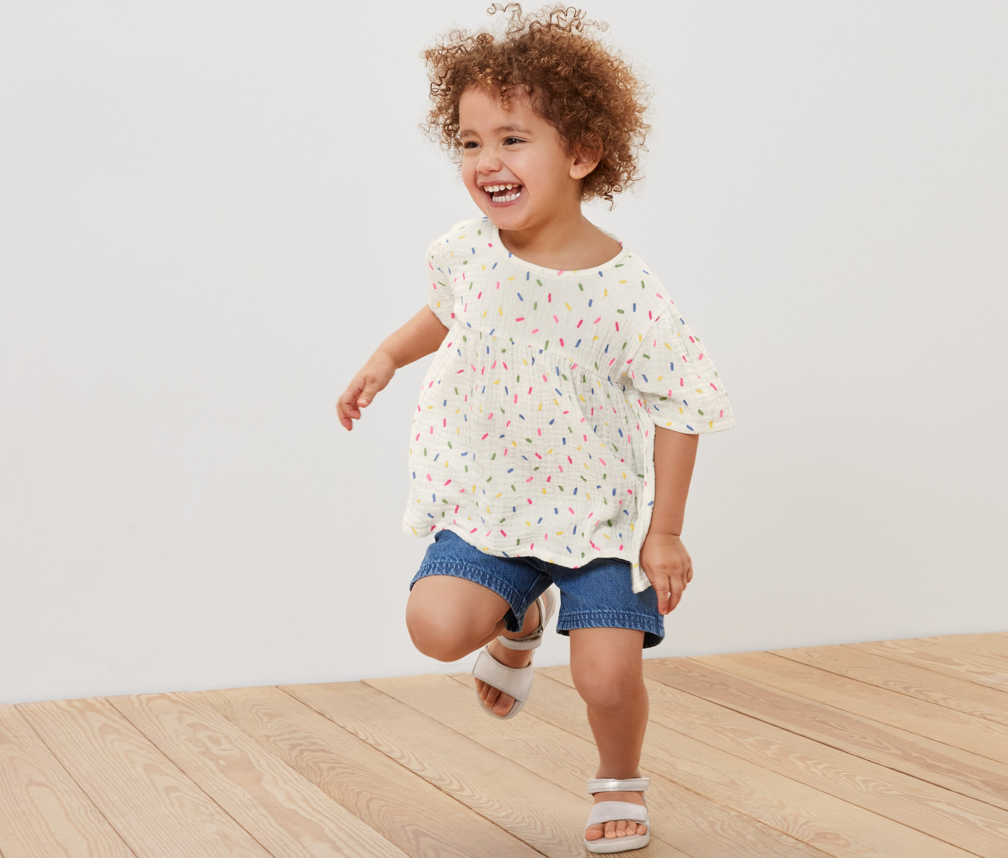 Fillette souriante aux cheveux bouclés courant et portant une blouse en mousseline pour enfant, un short en jean et des sandales.
