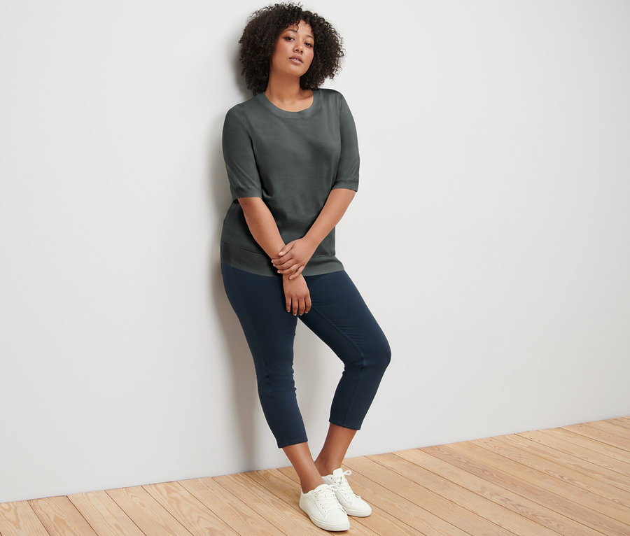 Une femme appuyée contre un mur porte un pull en maille fine vert, un pantalon bleu marine et des baskets blanches.