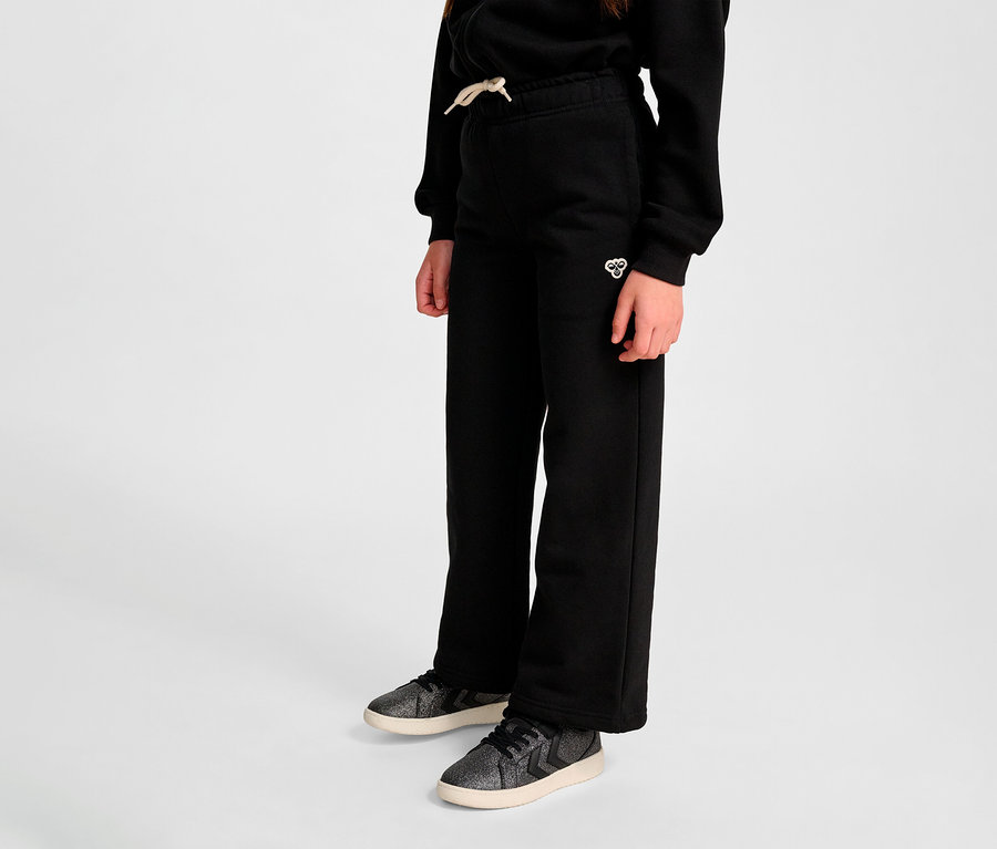 Gros plan d'une personne portant un pantalon de survêtement noir HUMMEL HMLJR Loose WL et des baskets sur un fond gris.