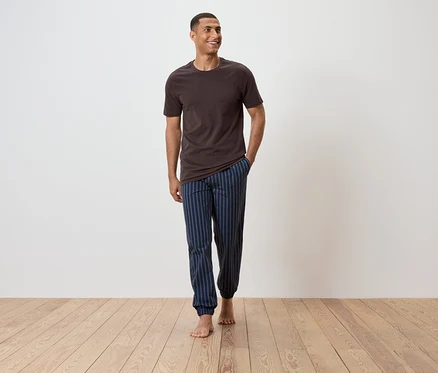 Un homme marche pieds nus sur un plancher en bois, portant un t-shirt marron et un pantalon de pyjama bleu rayé.