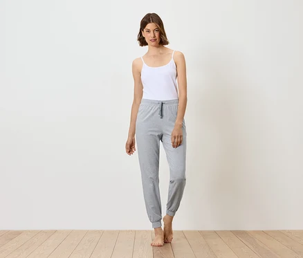 Femme debout pieds nus, portant un débardeur blanc et un pantalon de jogging gris.