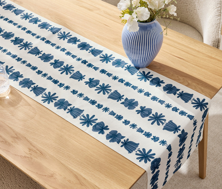 Table en bois beige avec un chemin de table bleu et blanc, un vase bleu avec des fleurs blanches et un verre.