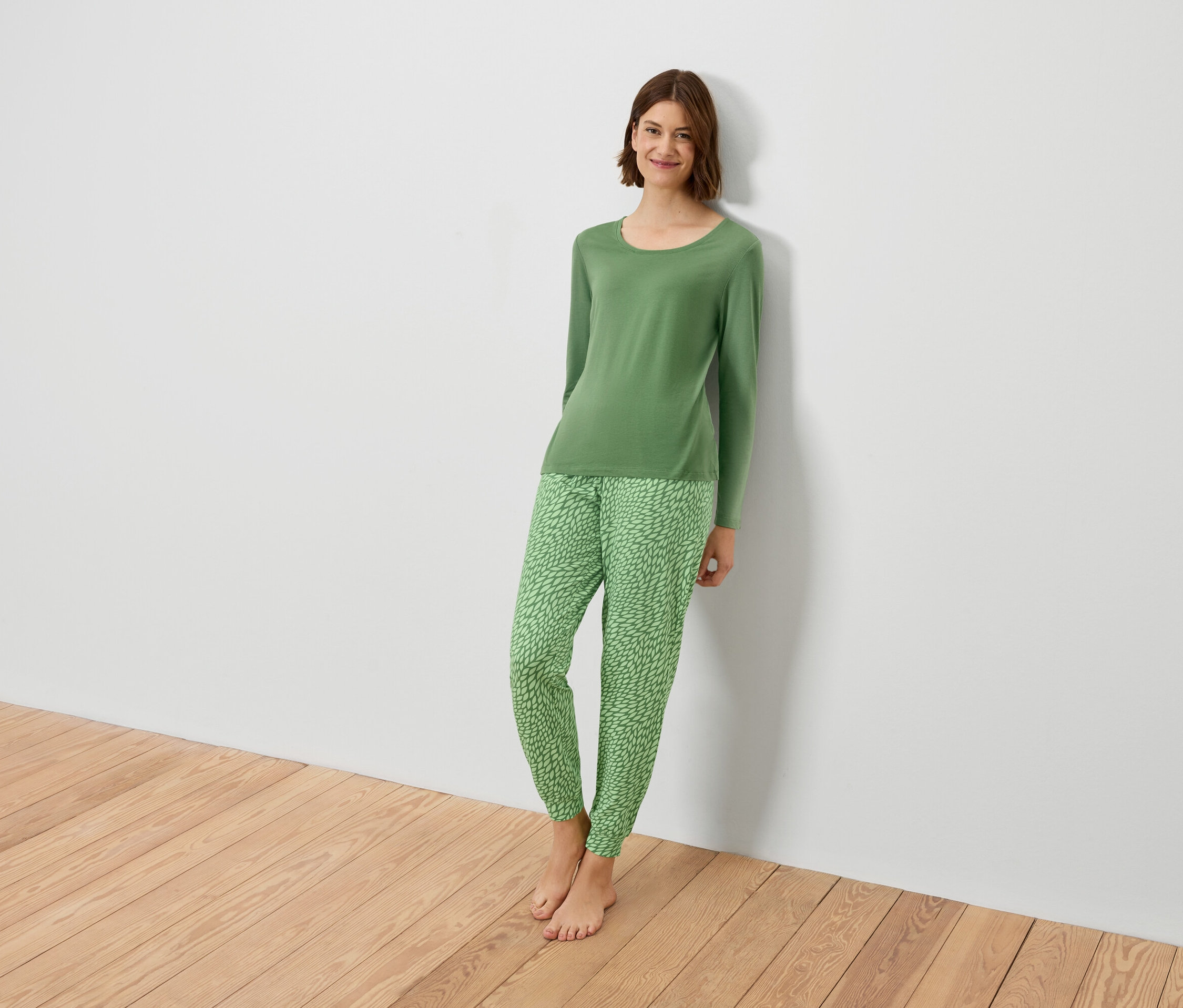 Une femme en pyjama vert est adossée à un mur blanc.