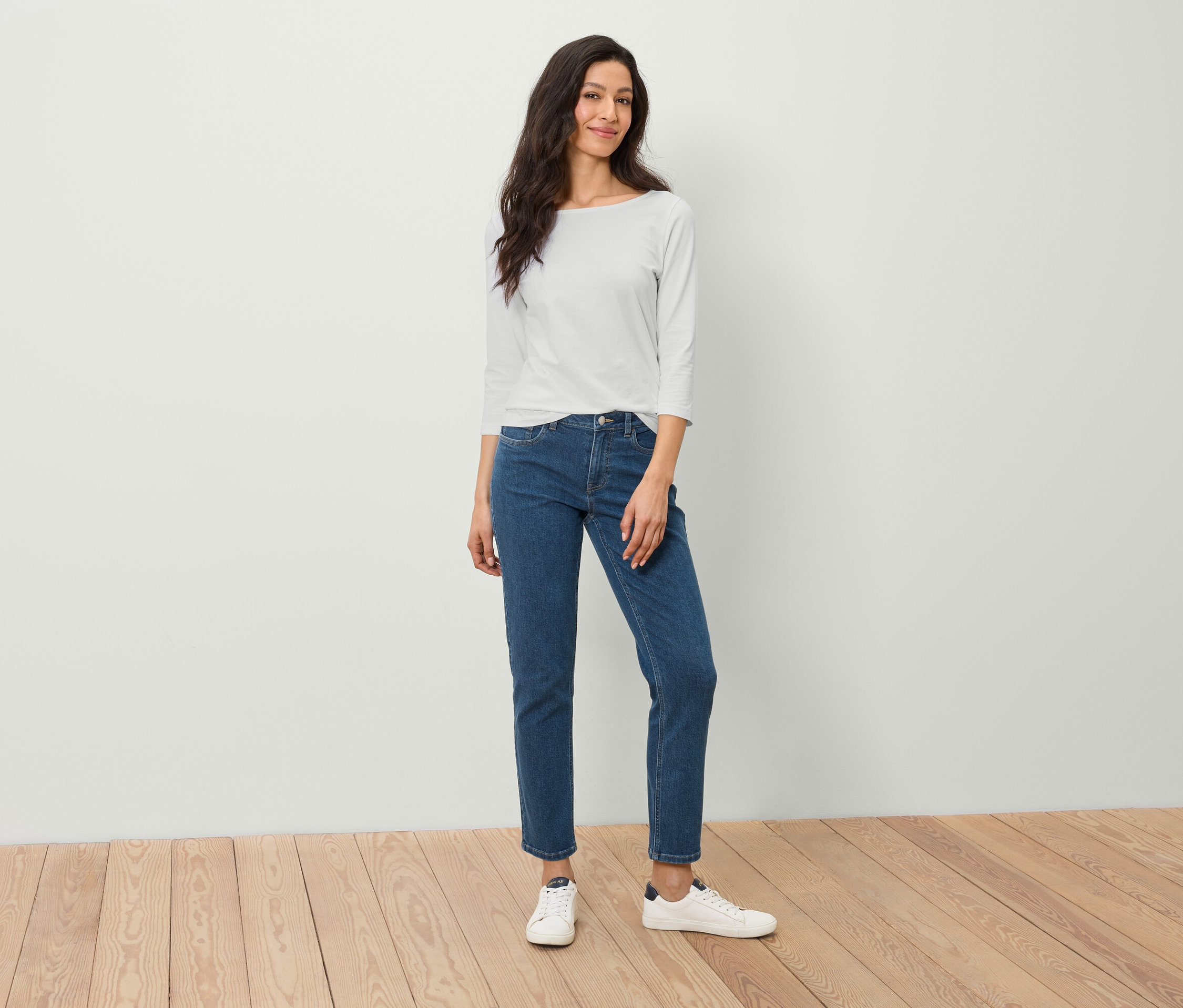 Une femme pose, portant un t-shirt blanc à manches trois-quarts, un jean bleu et des baskets blanches.