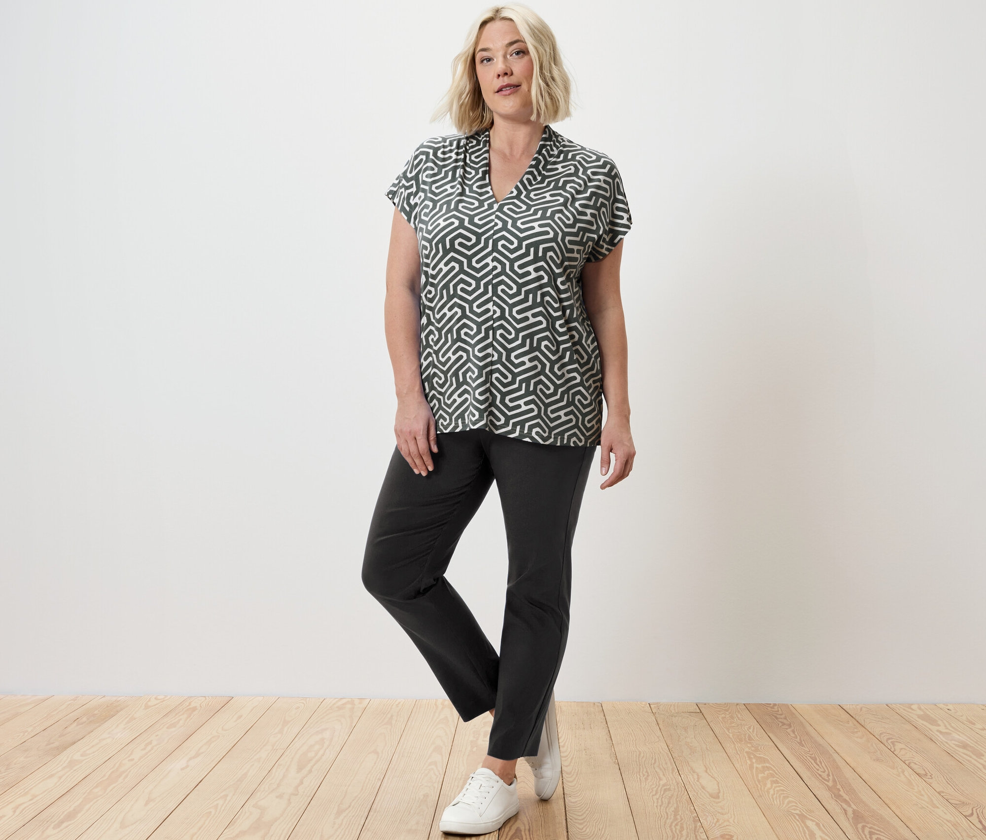 Une femme blonde pose en blouse verte à col en V et en pantalon stretch.