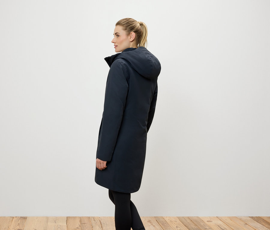 Vue latérale d'une femme aux cheveux blonds attachés en queue de cheval, portant un long manteau bleu foncé à capuche et des leggings noirs.