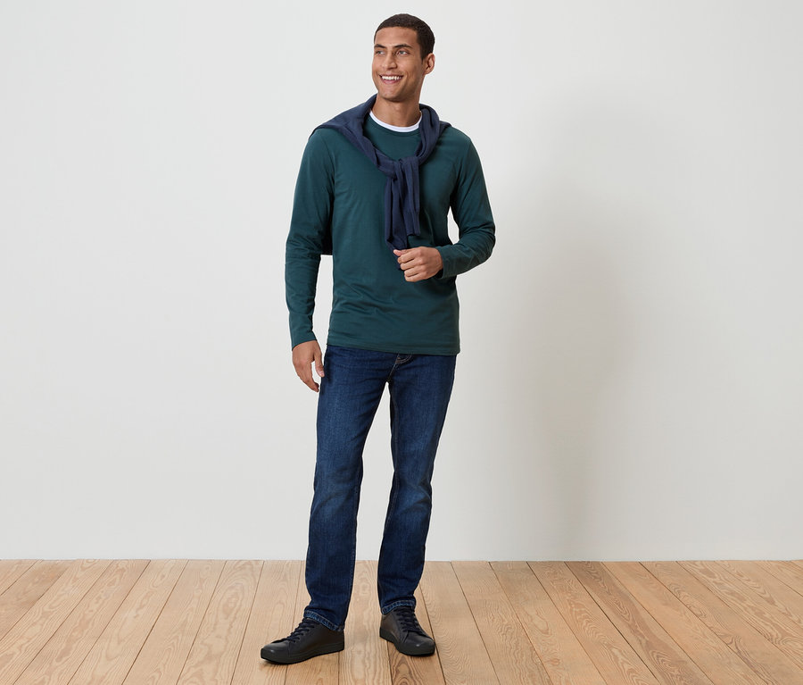 Un homme aux cheveux foncés et souriant se tient sur un plancher en bois. Il porte un t-shirt vert à manches longues, un jean bleu et des chaussures noires. Un pull bleu marine est drapé autour de son cou.