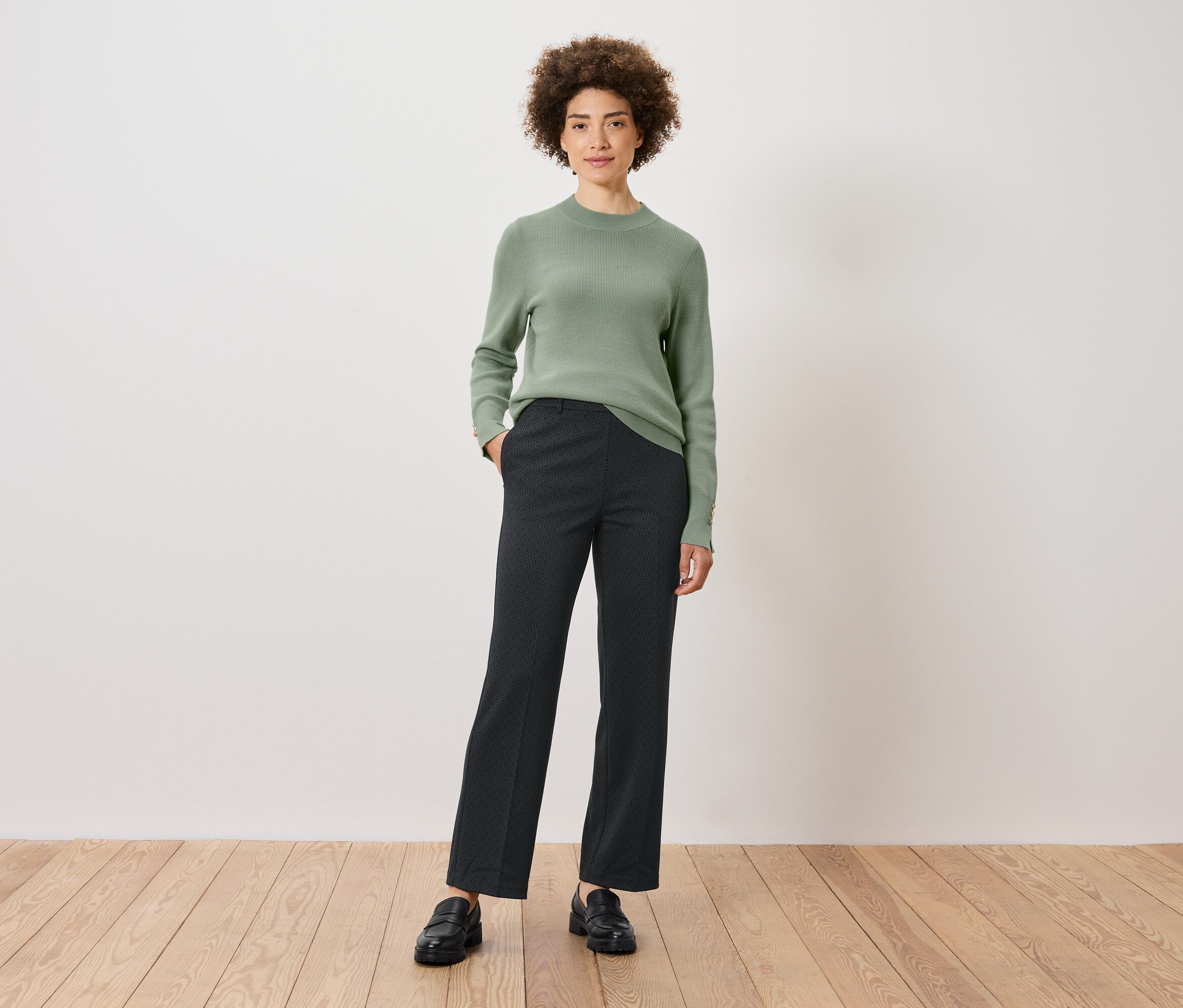 Une femme aux cheveux bouclés se tient debout, une main dans la poche de son pantalon. Elle porte un pull vert, un pantalon à motifs et des chaussures noires.