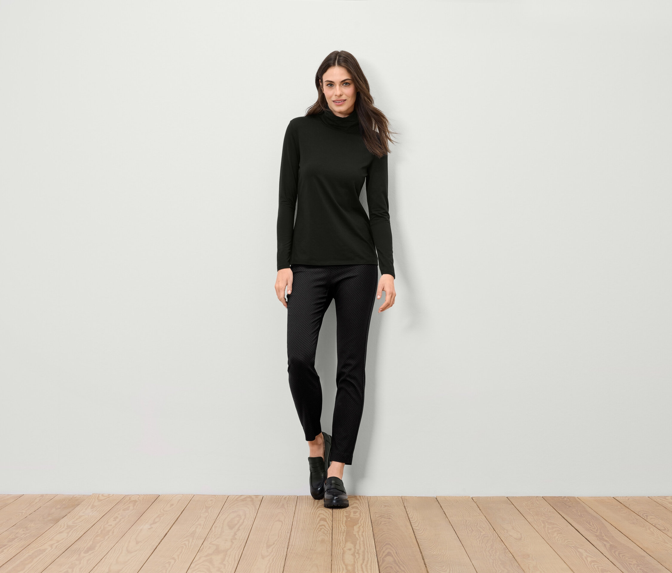 Une femme debout porte un pull à col roulé noir, un pantalon noir et des chaussures noires.