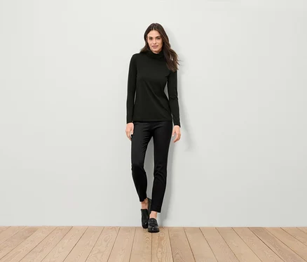Une femme debout porte un pull à col roulé noir, un pantalon noir et des chaussures noires.