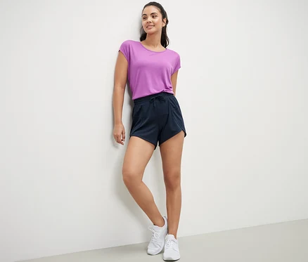 Une femme appuyée contre un mur porte un t-shirt violet et un short de sport bleu foncé.