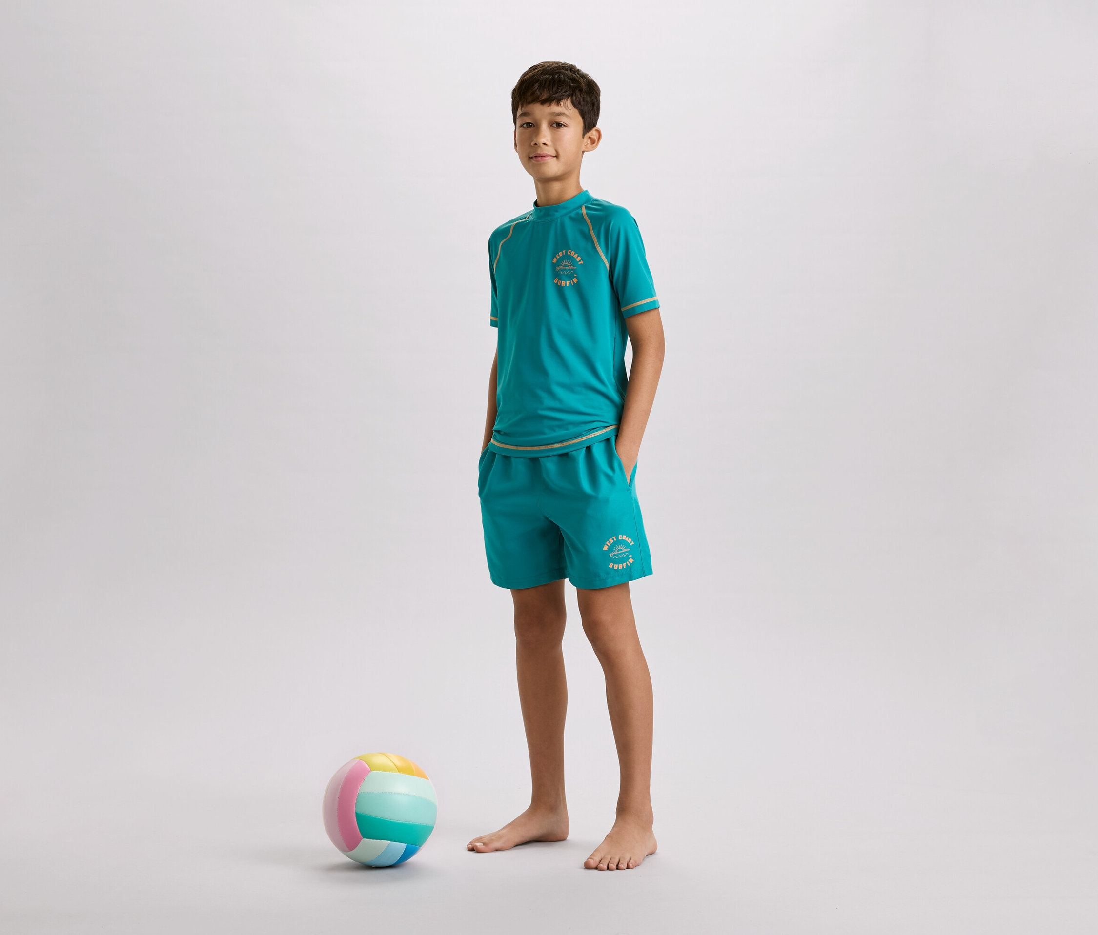 Un garçon aux pieds nus, vêtu d'un t-shirt turquoise et d'un short, se tient à côté d'un ballon coloré sur un fond gris.