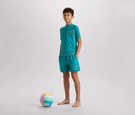 Un garçon aux pieds nus, vêtu d'un t-shirt turquoise et d'un short, se tient à côté d'un ballon coloré sur un fond gris.