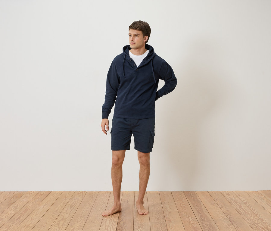 Un homme se tient pieds nus devant un mur blanc, portant un sweat à capuche bleu marine et un short.