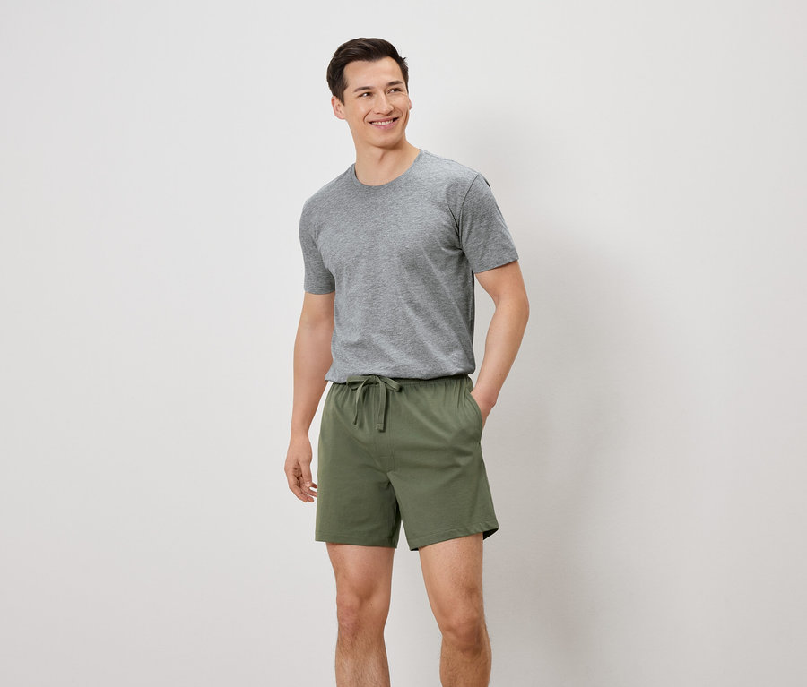Un homme portant un t-shirt gris et un short vert se tient devant un fond blanc.