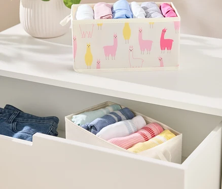 Sur une étagère blanche se trouve un récipient en tissu rempli de vêtements avec un motif de lama. En dessous se trouve un tiroir avec un autre récipient et des vêtements pliés.