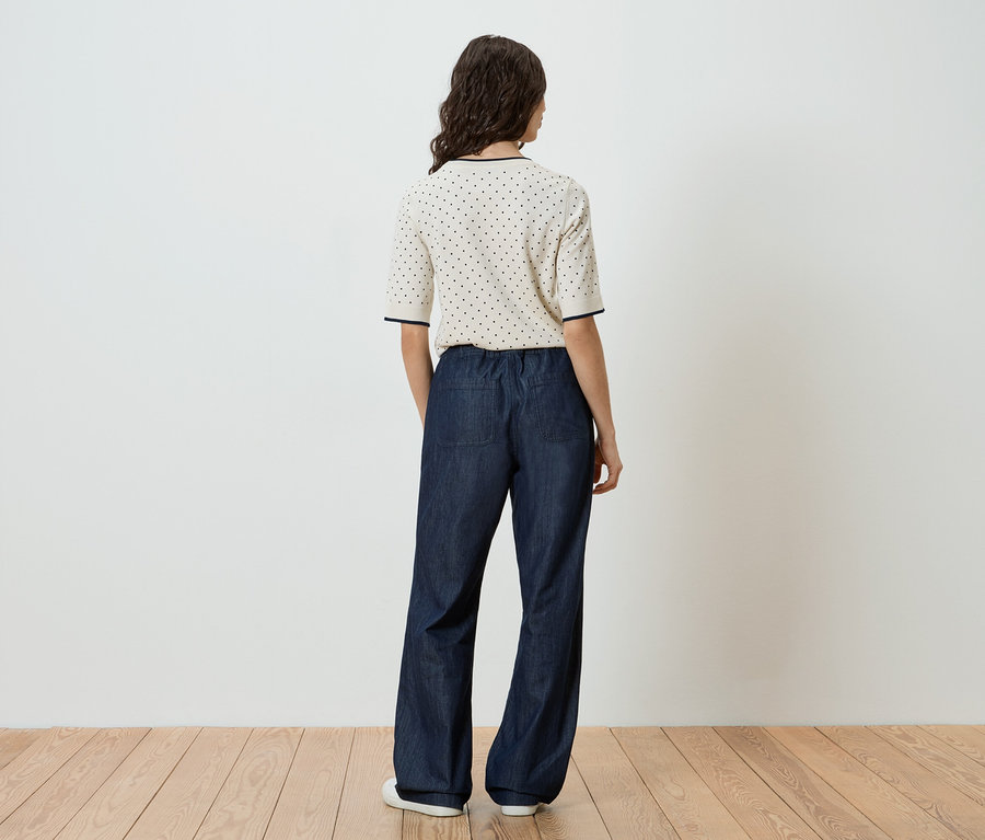 Vue arrière d'une femme aux cheveux ondulés et foncés portant un haut crème à manches courtes avec des pois bleu marine, un pantalon large bleu marine avec une taille élastique et des chaussures blanches.