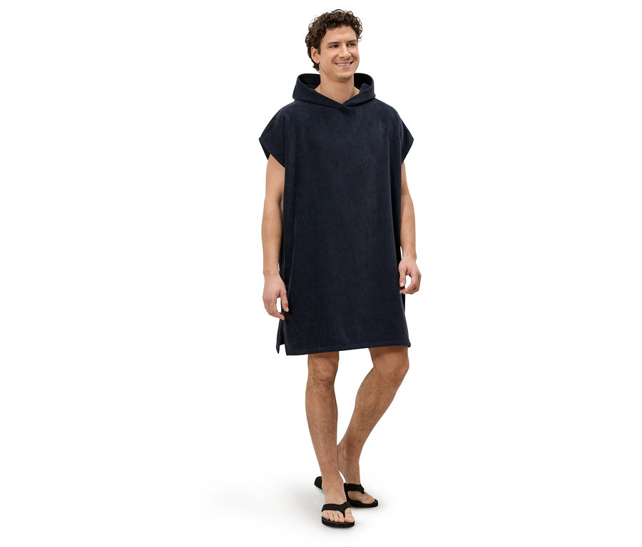 Un homme se tient pieds nus sur un fond blanc, vêtu d'un poncho à capuche bleu marine et de tongs noires.