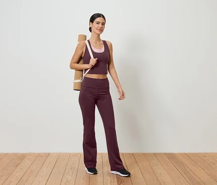 Une femme en tenue de sport avec un tapis de yoga sur l'épaule se tient sur un plancher en bois devant un mur blanc.