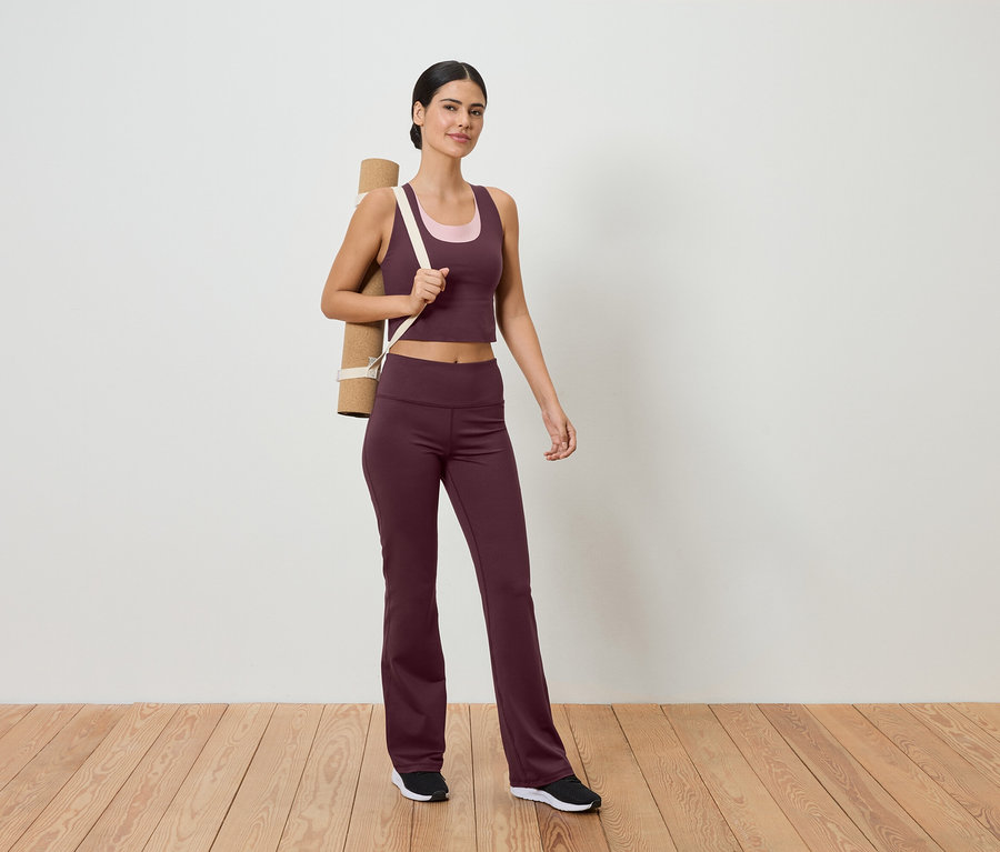 Une femme en tenue de sport avec un tapis de yoga sur l'épaule se tient sur un plancher en bois devant un mur blanc.