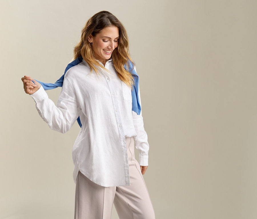 Une femme avec un pull bleu sur les épaules porte une blouse longue en lin blanche.