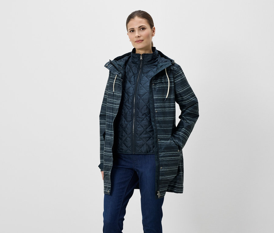 Femme portant un jean, un gilet matelassé et un imperméable rayé à capuche. La veste a des poches et un cordon clair.