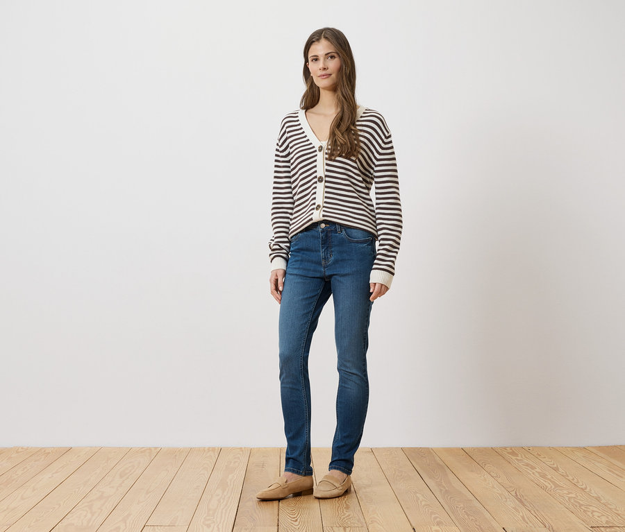 Une femme en jean bleu, cardigan rayé et chaussures beiges se tient devant un mur blanc sur un plancher en bois.