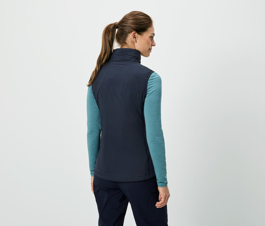 Vue de dos d'une femme avec une queue de cheval portant un gilet bleu marine sur une chemise turquoise à manches longues et un pantalon bleu marine.