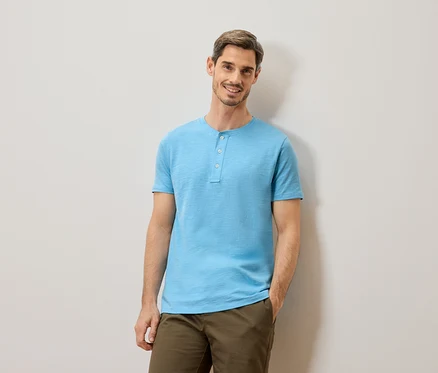 Un homme aux cheveux bruns courts et à la barbe, portant un t-shirt bleu clair à boutons et un pantalon kaki, se tient devant un mur gris clair.