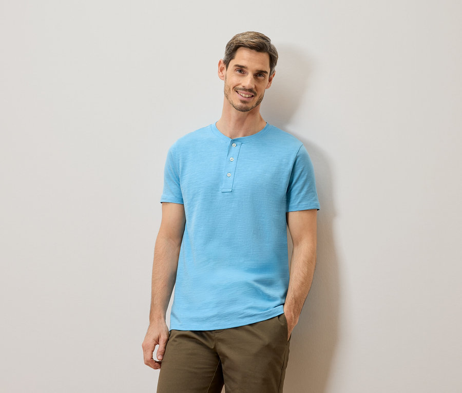 Un homme aux cheveux bruns courts et à la barbe, portant un t-shirt bleu clair à boutons et un pantalon kaki, se tient devant un mur gris clair.