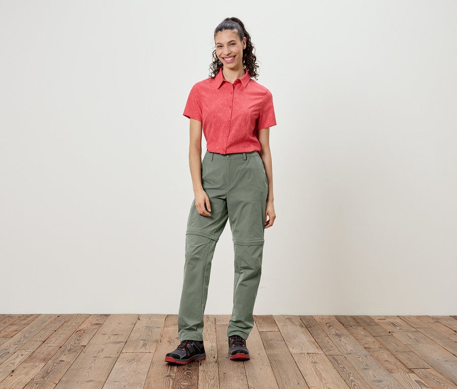 Une femme se tient devant un mur, portant une chemise rouge à manches courtes et un pantalon vert.