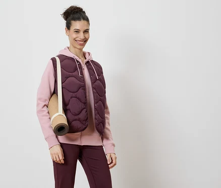 Une femme porte un gilet matelassé bordeaux sur un sweat à capuche rose et un legging bordeaux, avec un tapis de yoga sur l'épaule.