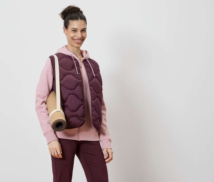 Une femme porte un gilet matelassé bordeaux sur un sweat à capuche rose et un legging bordeaux, avec un tapis de yoga sur l'épaule.