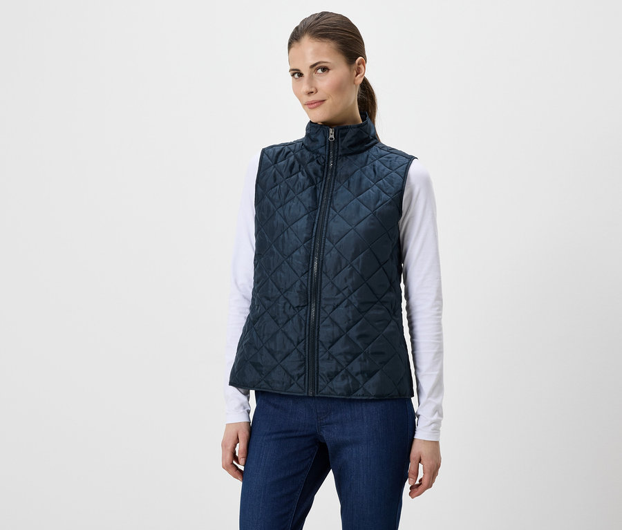 Une femme portant une queue de cheval porte un gilet matelassé bleu marine sur un t-shirt blanc à manches longues et un jean bleu.