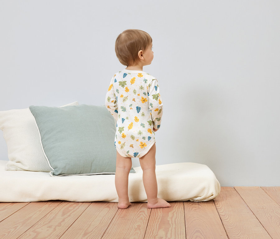 Un jeune enfant en body à manches longues avec un motif de feuilles se tient sur un plancher en bois à côté de coussins et d'un matelas.