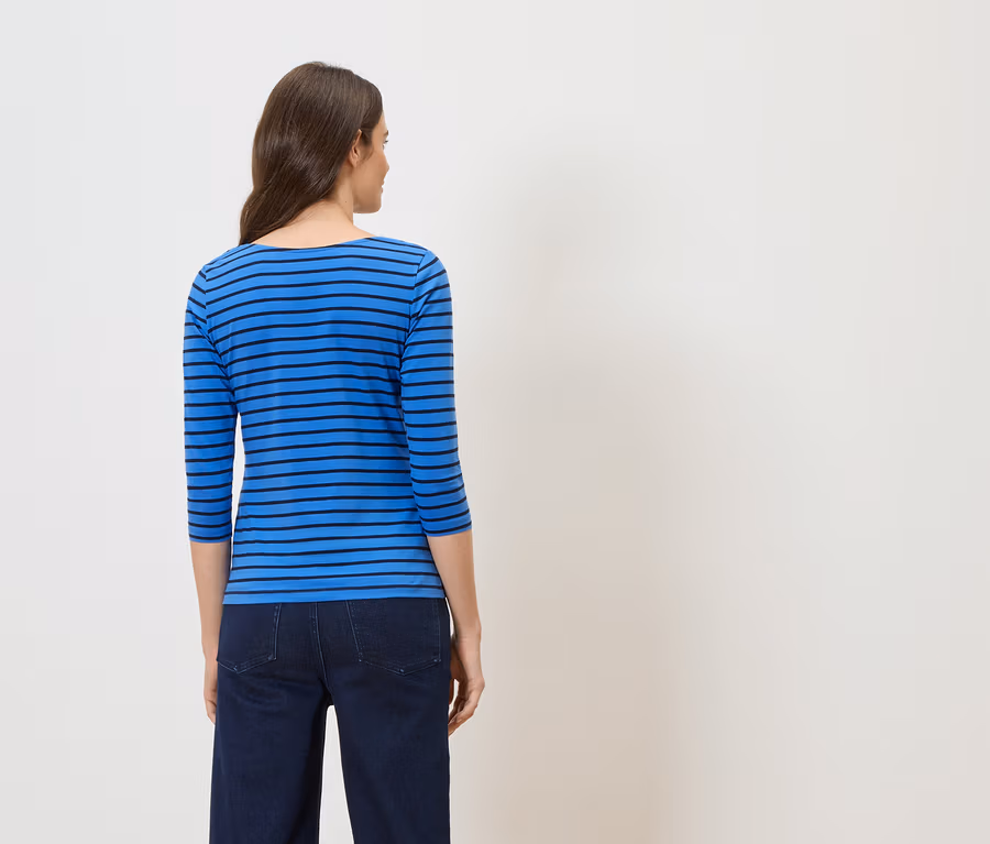 Vue de dos d'une femme aux cheveux bruns portant un haut rayé bleu et noir et un jean foncé sur fond blanc.
