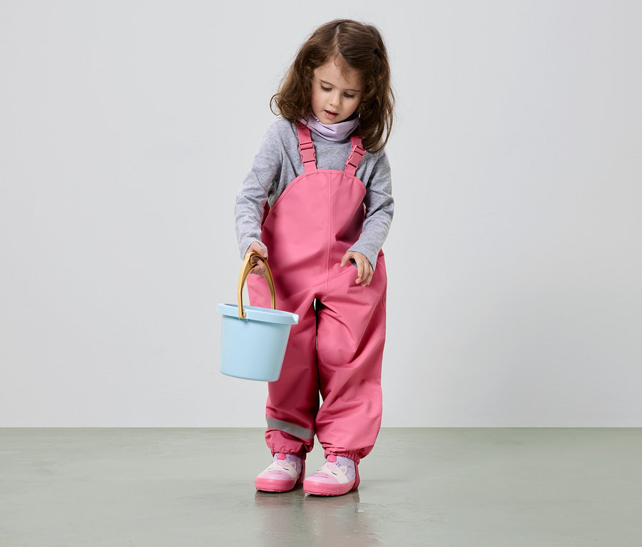 Une fillette aux cheveux bruns bouclés tient un seau bleu clair, porte une salopette rose et des chaussures roses.