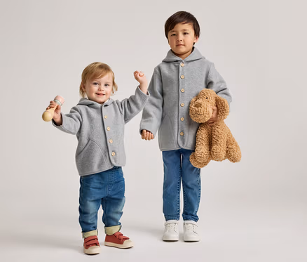 Un tout-petit et un enfant plus âgé se tiennent côte à côte. Tous deux portent des vestes grises à capuche avec des boutons en bois et des jeans bleus. Le tout-petit à gauche tient un hochet en bois et porte des chaussures rouges. L'enfant plus âgé à droite tient un chien en peluche marron et porte des chaussures blanches.