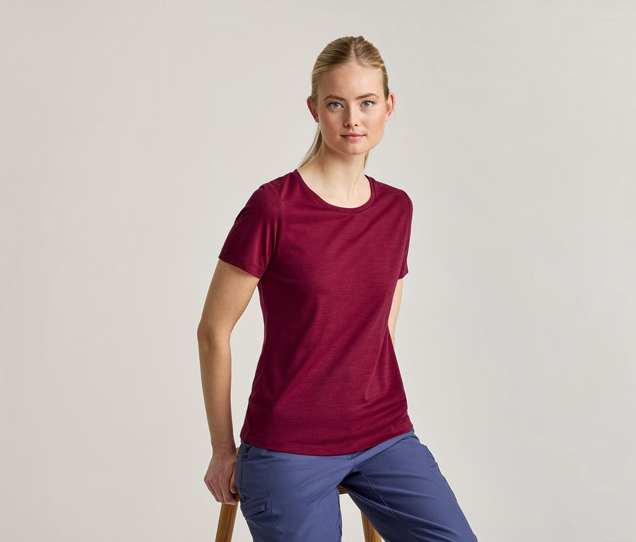 Une femme aux cheveux blonds attachés, vêtue d'un t-shirt bordeaux à manches courtes et d'un pantalon bleu, est assise sur un tabouret en bois et regarde l'appareil photo.
