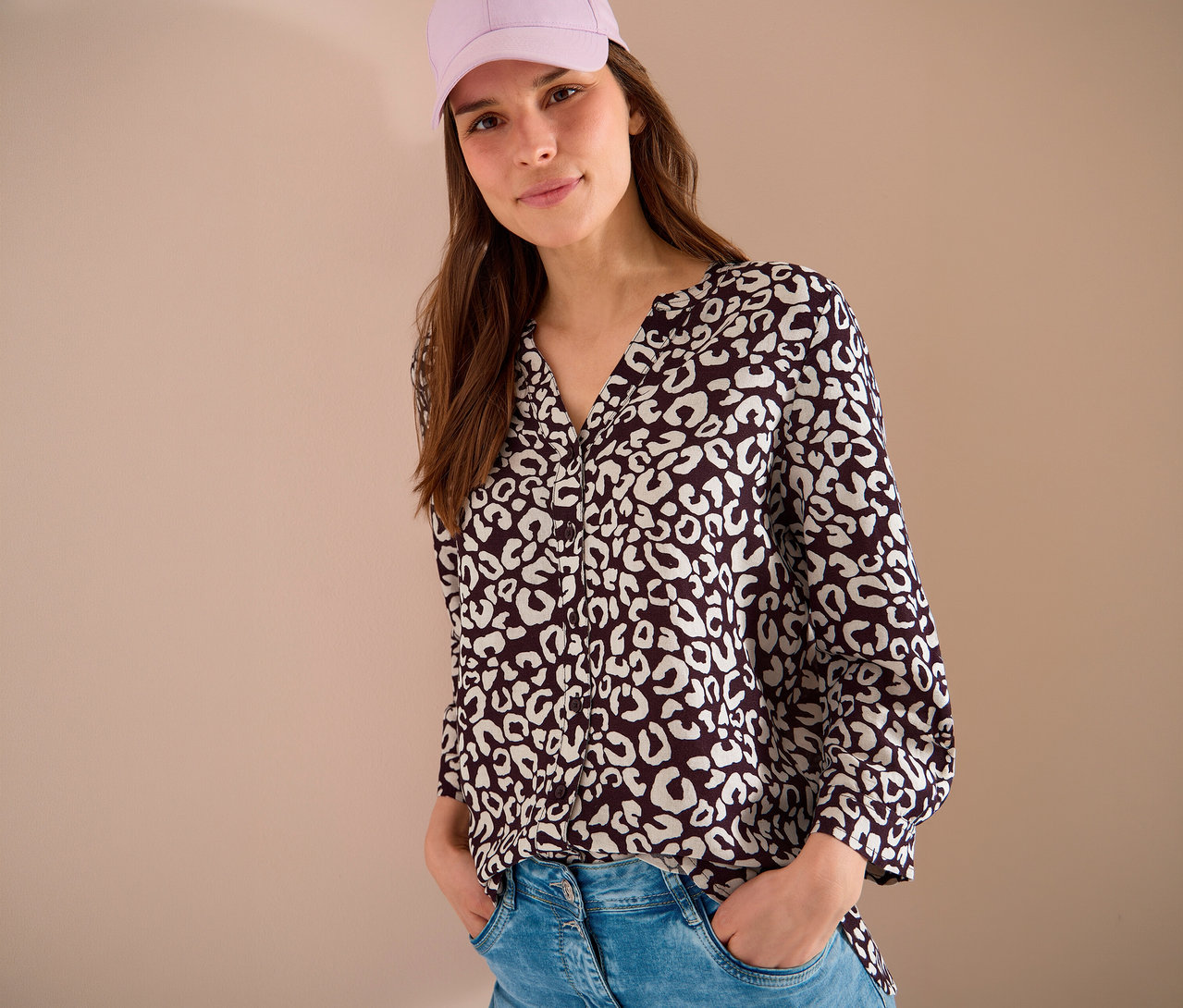 Une femme portant une casquette rose et un chemisier à motifs, les mains dans les poches de son jean.