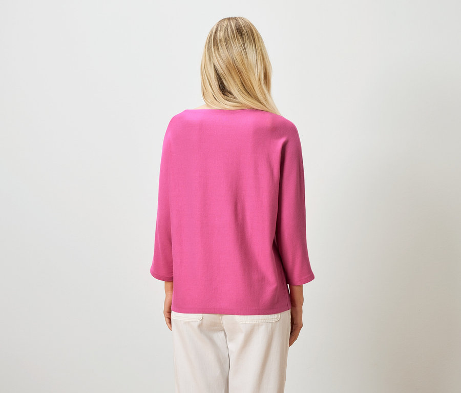 Vue de dos d'une femme blonde portant un haut rose et un pantalon clair sur un fond blanc.