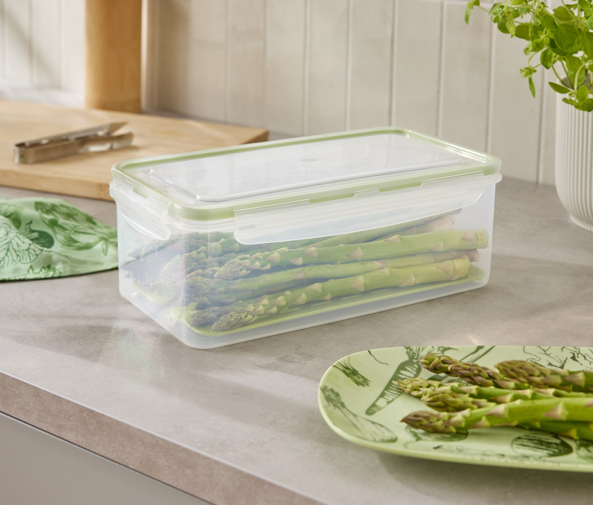 Sur un plan de travail de cuisine se trouvent un récipient contenant des asperges, une assiette d'asperges et une plante.