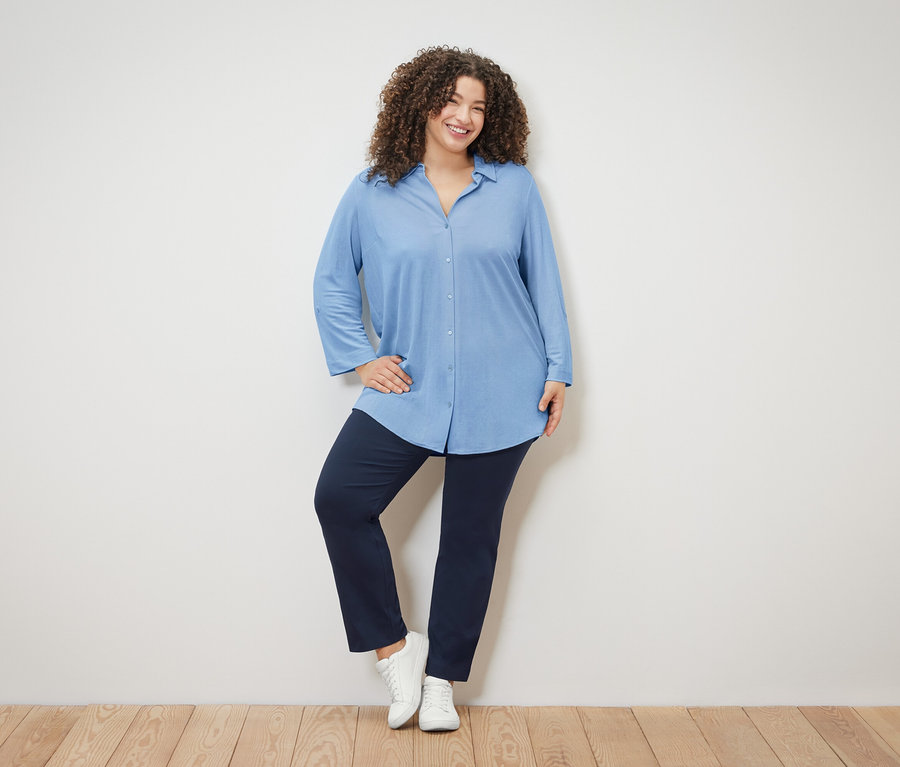 Une femme aux cheveux bouclés porte un chemisier bleu clair, un pantalon bleu marine et des chaussures blanches.
