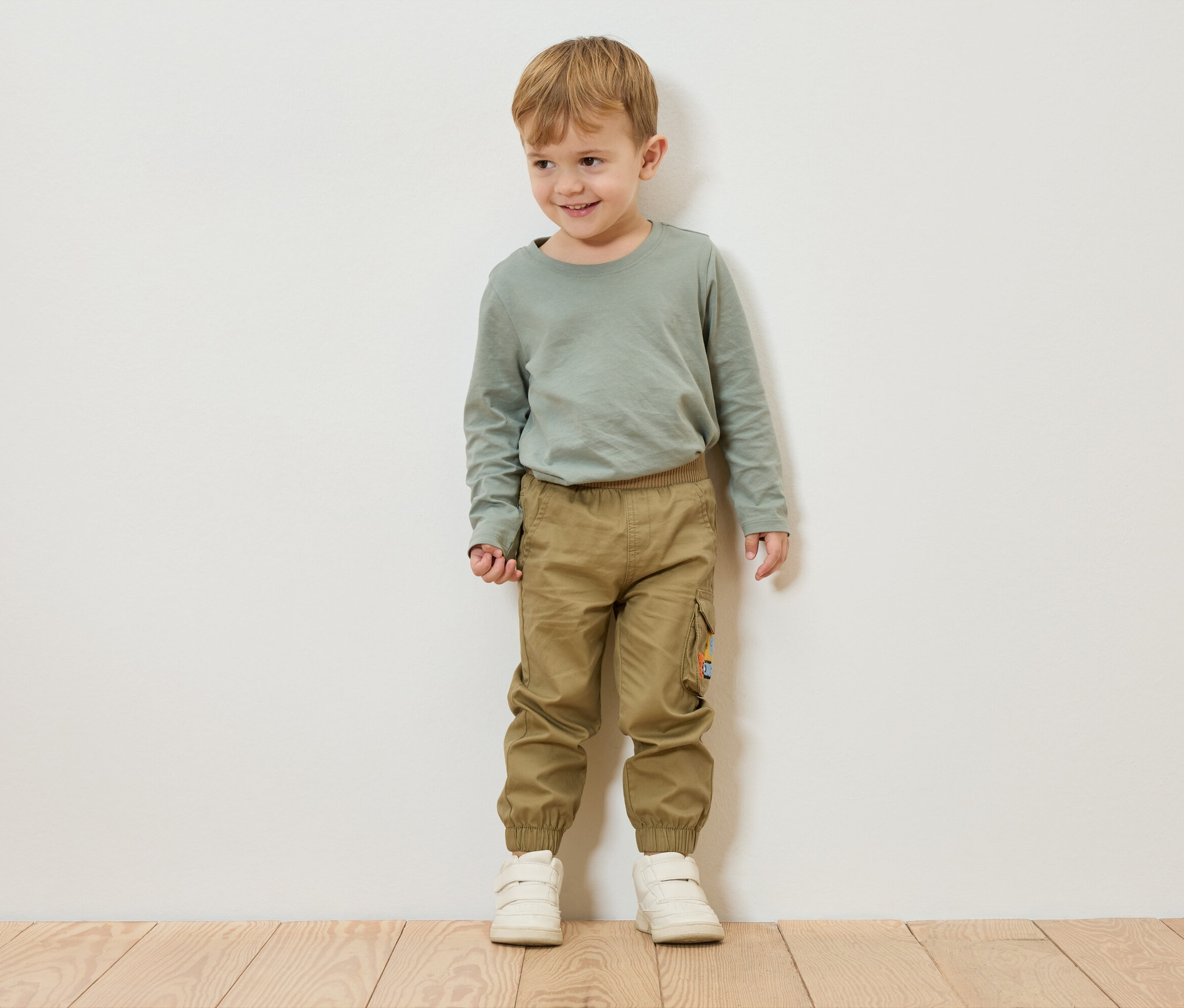 Un garçon aux cheveux blonds se tient devant un mur blanc, portant un t-shirt vert à manches longues, un pantalon kaki et des chaussures blanches.