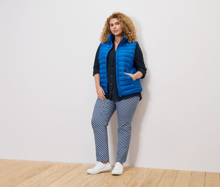 Une femme aux cheveux bouclés portant un gilet bleu, une chemise foncée et un pantalon à motifs se tient contre un mur blanc.