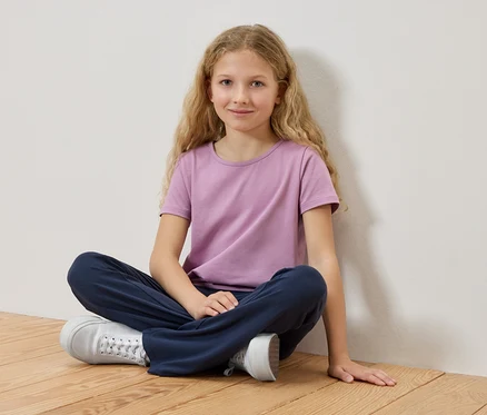 Une fille aux cheveux blonds est assise les jambes croisées sur un plancher en bois, portant un t-shirt violet et un pantalon bleu.
